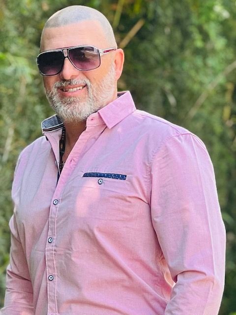Man wearing sunglasses and a pink shirt, smiling outdoors with foliage in the background.